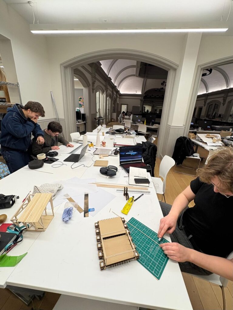 Three people work on architectural models at tables in a spacious, well-lit studio with high ceilings and arched doorways. Drafting tools, laptops, and scale models are scattered across the tables.