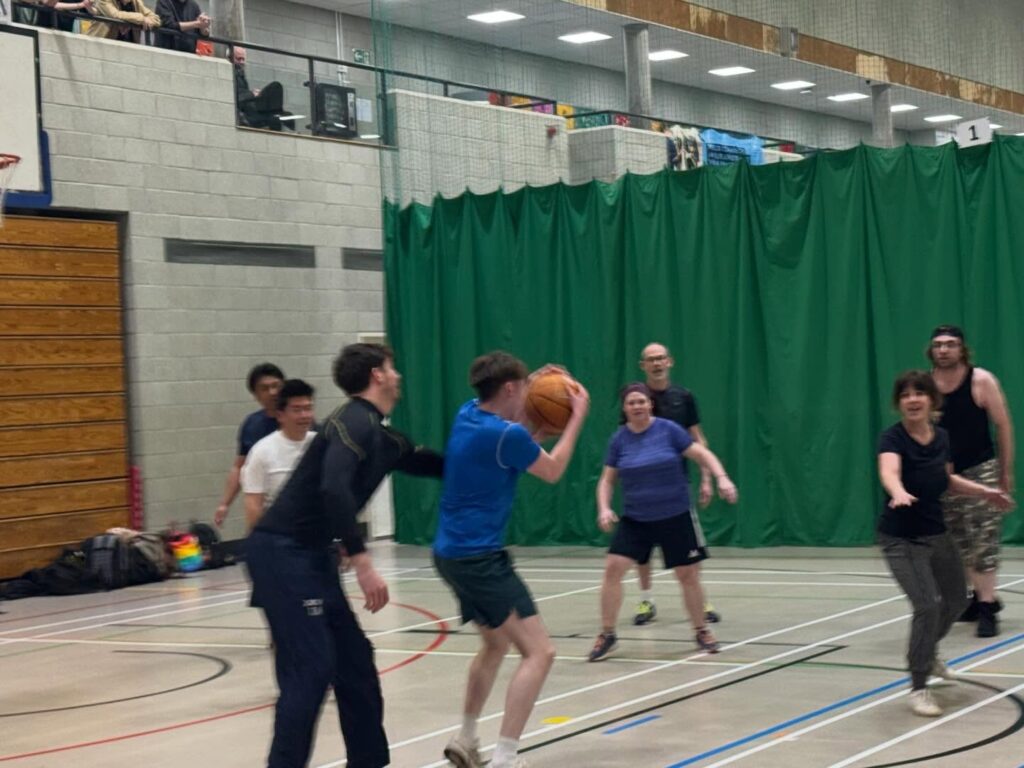 A group of adults play basketball in an indoor gym. One player in blue prepares to shoot while others, dressed casually, defend or watch. A green curtain and concrete walls are visible in the background.