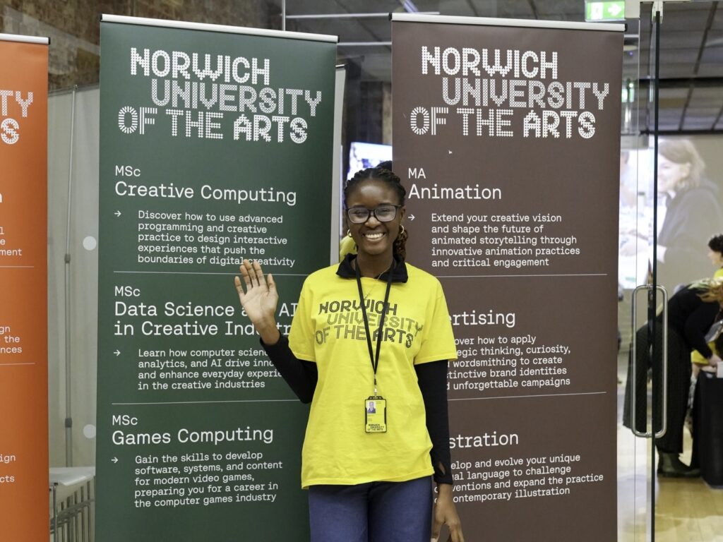 A smiling student wearing a yellow "Norwich University of the Arts" t-shirt and lanyard stands indoors, waving in front of banners displaying creative computing, games computing, animation, and other course details.