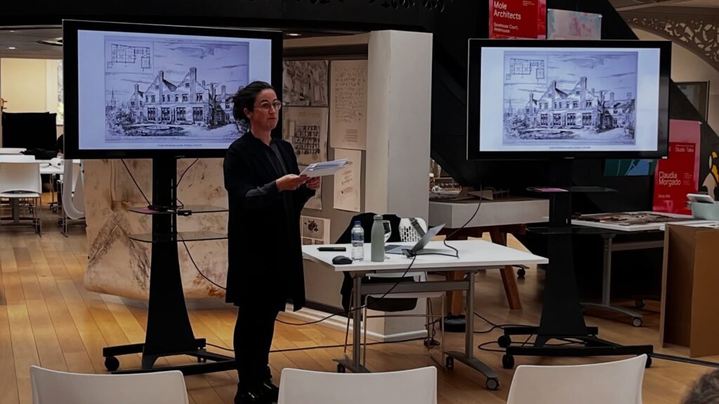 A person stands in front of two screens displaying architectural sketches of a building, holding papers and speaking in a modern, open space with desks, chairs, and architectural displays.