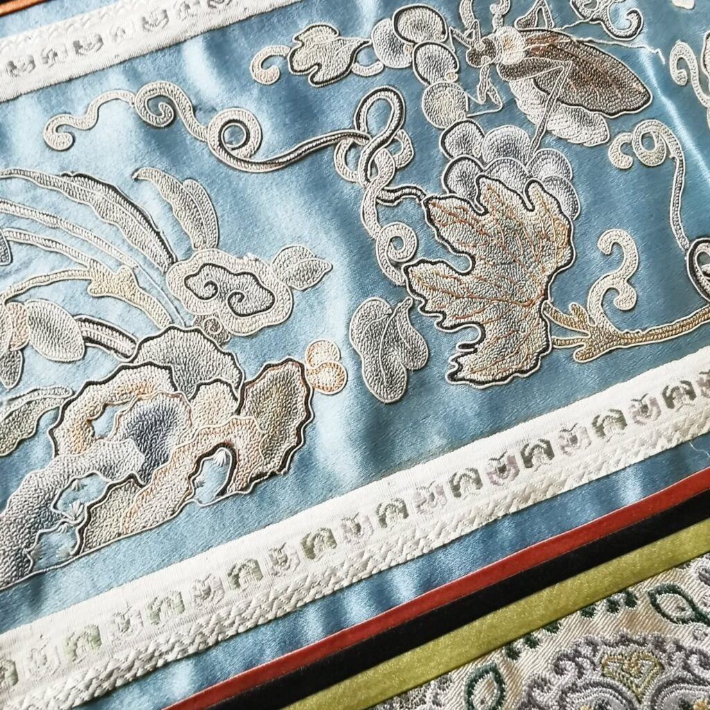 Close-up of ornate blue fabric with intricate embroidered floral and leaf patterns in silver and white, bordered by decorative bands in white and red. The design has a luxurious and traditional appearance.
