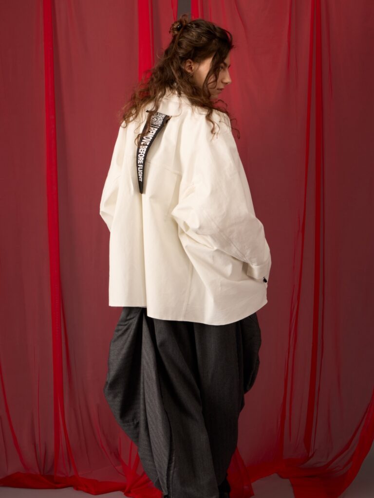 A person stands with their back turned, wearing a loose white shirt with black and white graphic detail on the back, and wide, dark gray pants. Red sheer fabric hangs in the background.