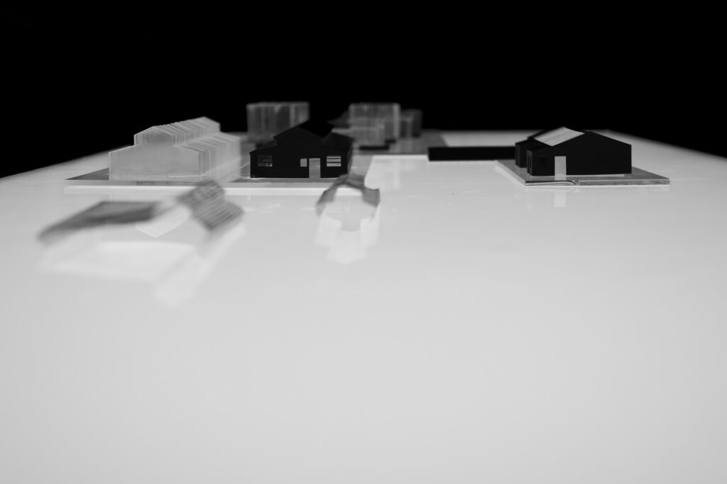 A black and white photo of a minimalistic architectural model featuring small, translucent and opaque geometric buildings arranged on a glossy white surface against a black background.