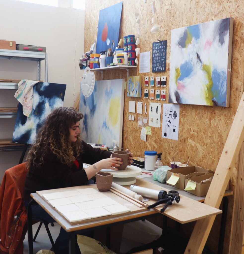 A woman with curly hair works with clay at a table in an art studio, surrounded by abstract paintings, art supplies, and tools hanging on a wooden wall.