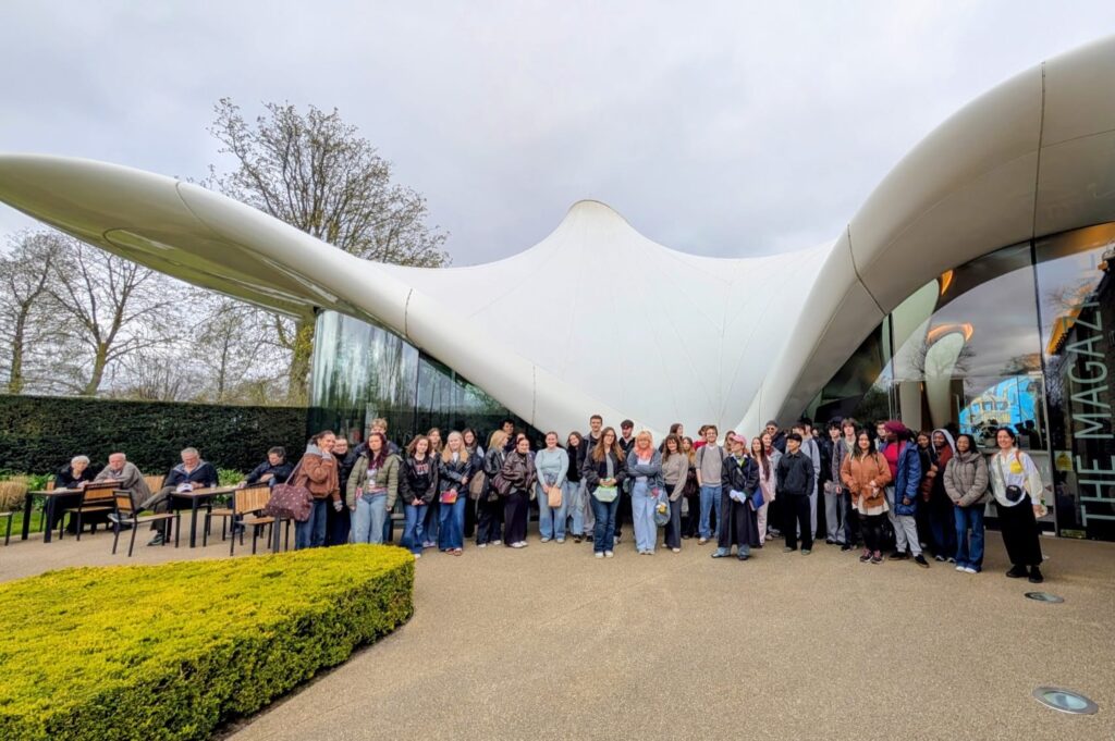 A large group of people stands in front of a modern, white, tent-like building with curved architecture. Some people sit at tables on the left, while trees and cloudy skies are seen in the background.