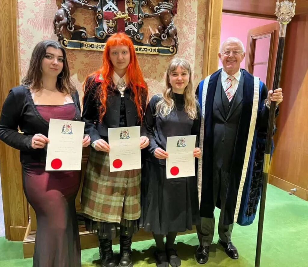 Three women holding certificates stand next to a smiling man in ceremonial robes holding a staff. They are indoors, in front of a decorative wooden wall with a crest.