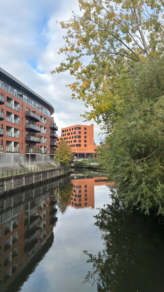 A calm river reflects modern red-brick apartment buildings and green trees under a partly cloudy sky. The scene is peaceful with no people visible.