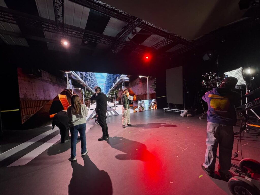People stand and work on a film set with a large digital backdrop showing an urban scene. Studio lights and cameras are visible, and crew members prepare for filming in a spacious, dark room.
