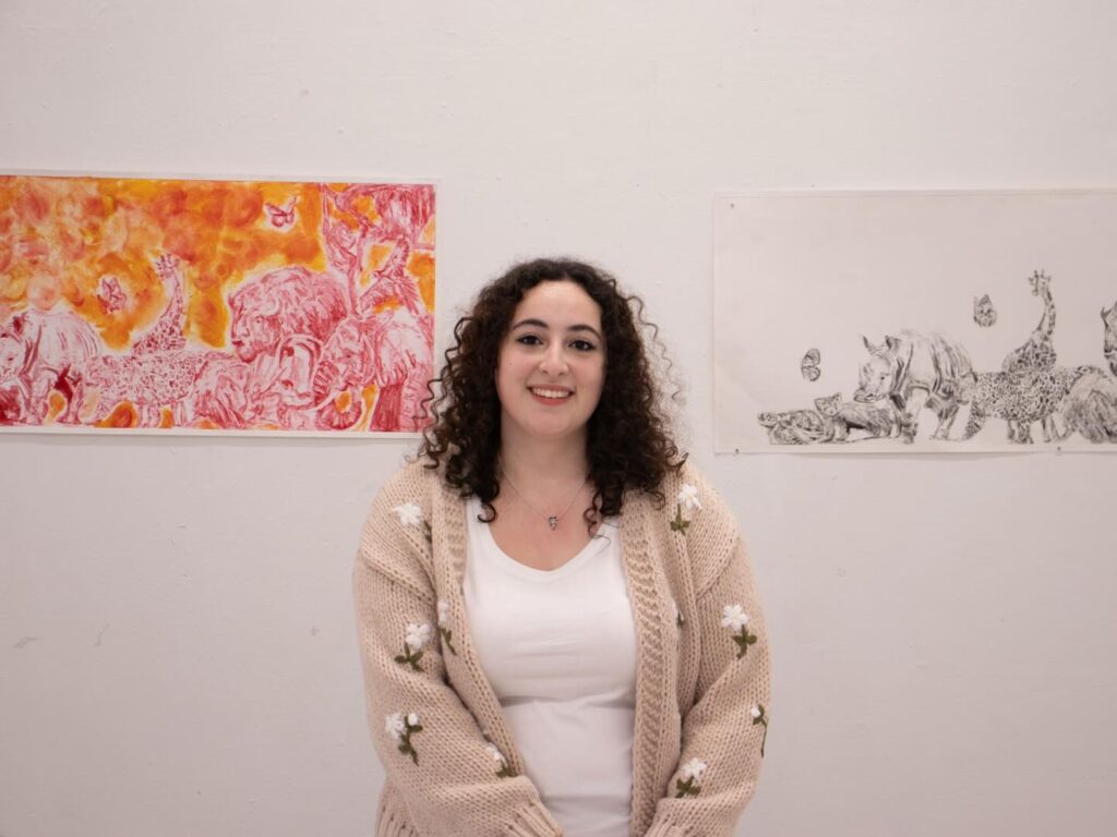 A woman with curly hair, wearing a beige sweater with flower patterns and a white shirt, stands smiling in front of two animal-themed artworks mounted on a white wall.