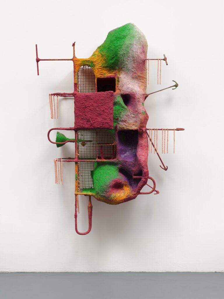 A colorful abstract sculpture mounted on a wall, featuring textured, rock-like surfaces in green, pink, yellow, and purple, intersected by red metal rods and wire mesh grids.