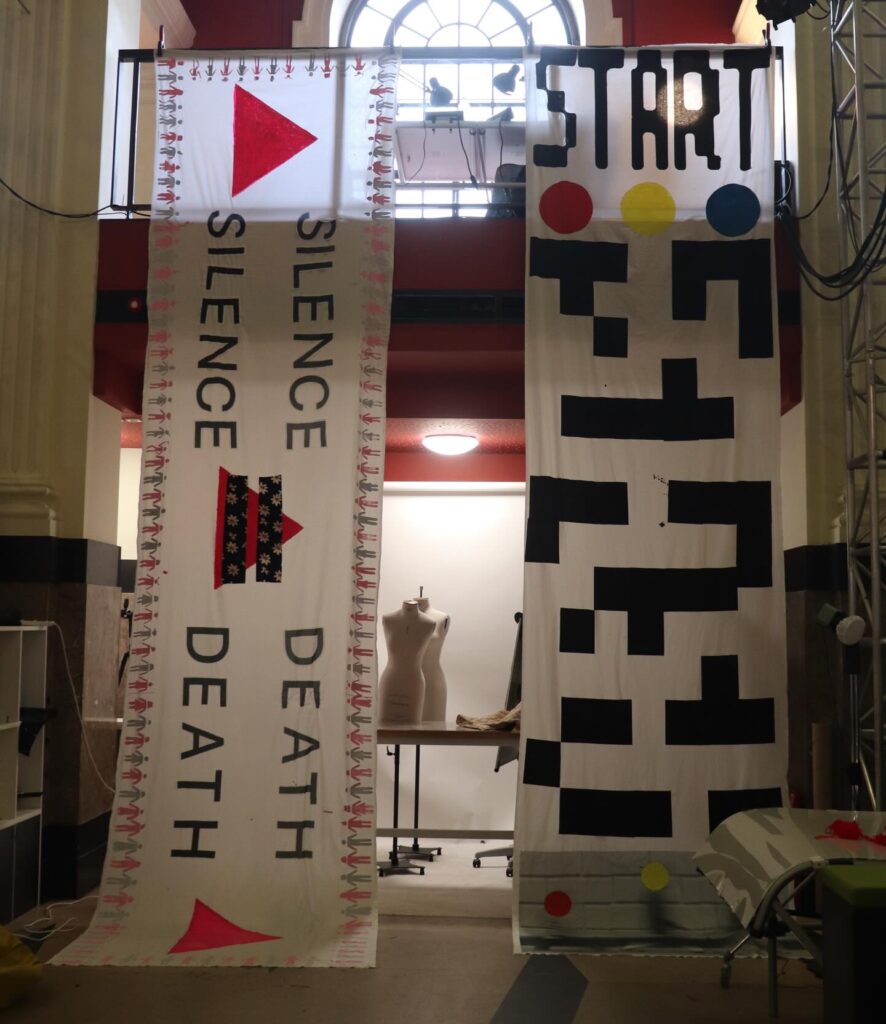 Two large banners hang indoors. The left banner reads "SILENCE = DEATH" with pink triangles and smaller black and red details. The right banner says "START" at the top with abstract black, yellow, blue, and red shapes. A dress form stands in the background.