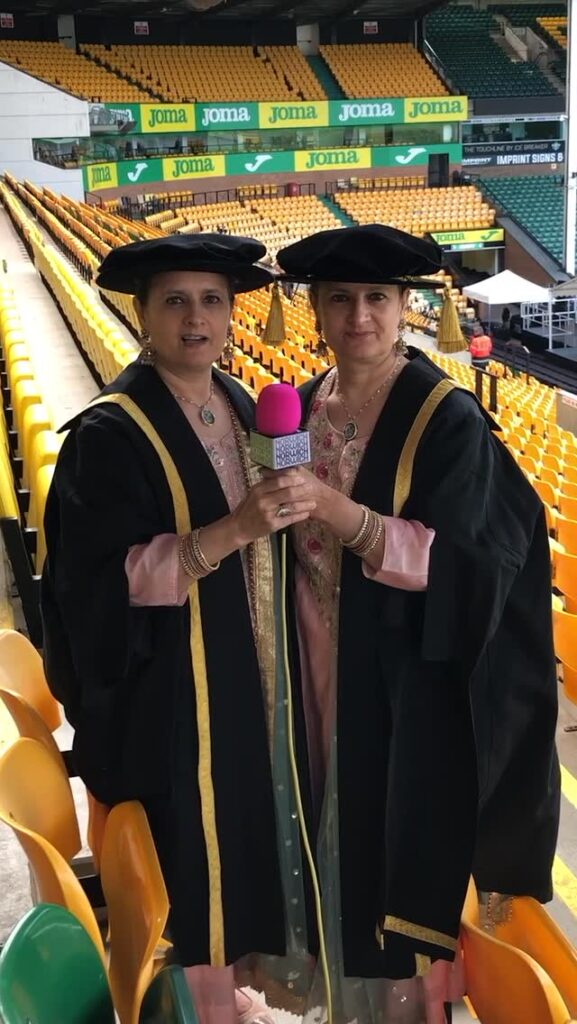 Two women in black and gold graduation gowns and caps stand in stadium seating, holding a pink microphone together. Yellow and green seats fill the background.
