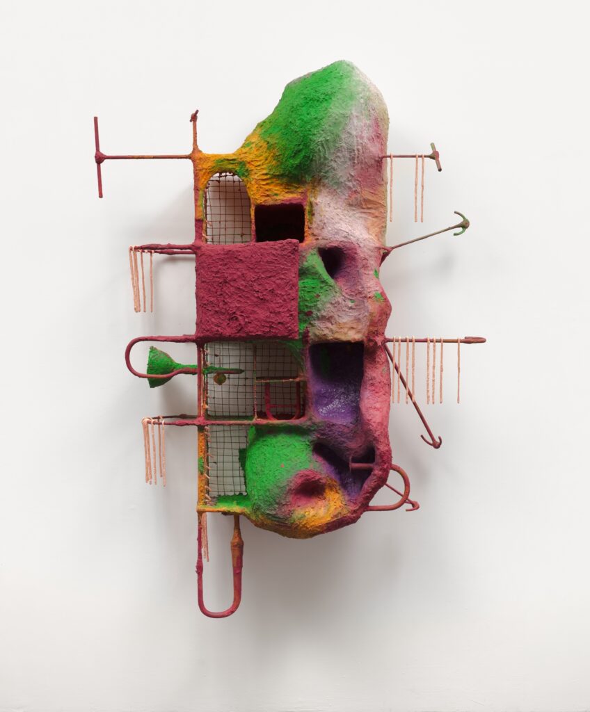 A colorful abstract sculpture featuring a lumpy, textured form with pink, green, orange, and yellow hues, intersected by gridded metal squares and rods, attached to a white wall.