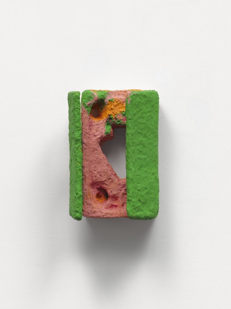 A small abstract sculpture mounted on a white wall, featuring textured pink and orange surfaces with holes, bordered by rough green edges, forming an open rectangular shape.