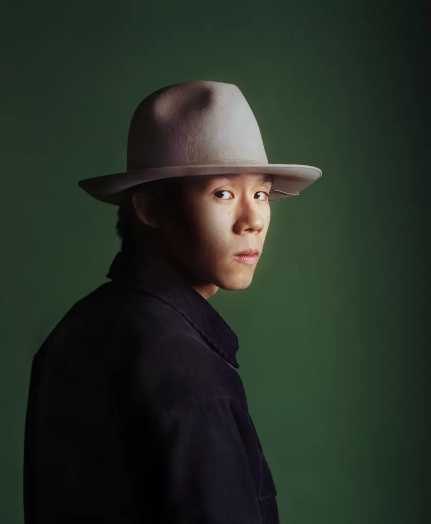 A person wearing a light-colored wide-brimmed hat and a dark jacket looks over their shoulder against a plain green background.