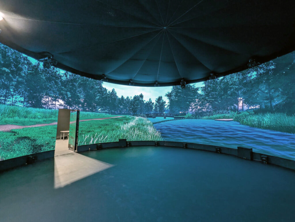 A circular room with a panoramic digital screen displaying a realistic outdoor scene featuring grass, trees, and water. An open door reveals a different floor surface leading outside the display area.
