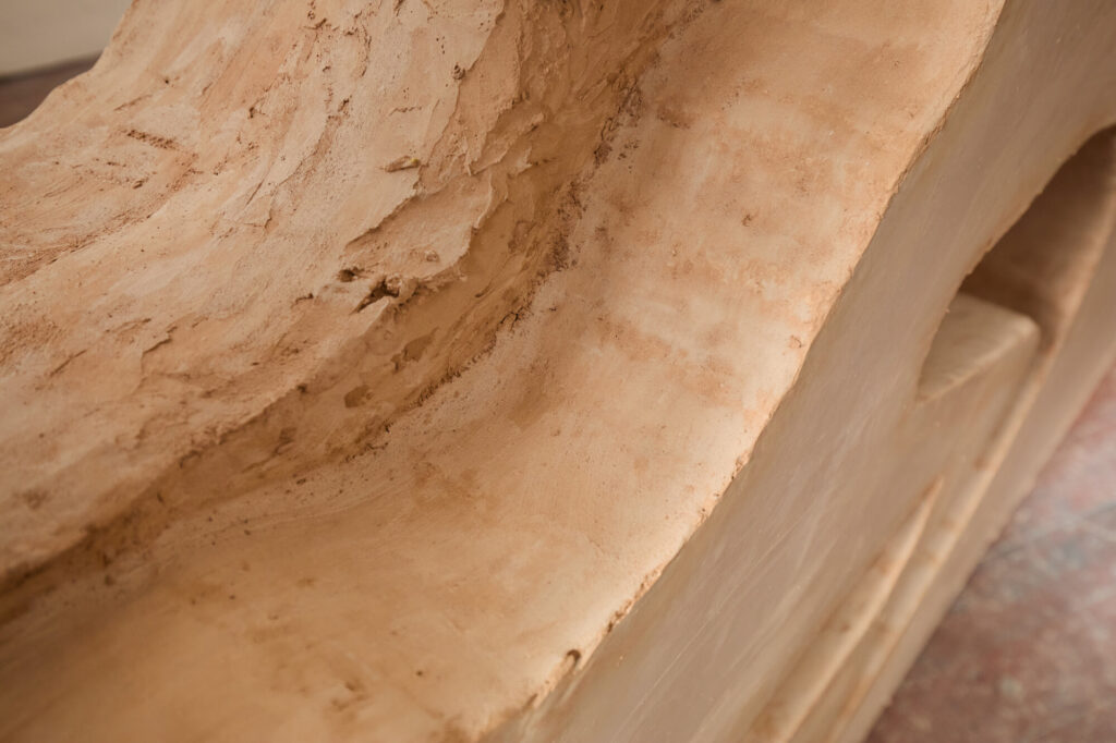 Close-up of a smooth, beige clay sculpture with curved and textured surfaces, showing natural grooves and hand-shaped details. The material appears unglazed and has an earthy, organic look.