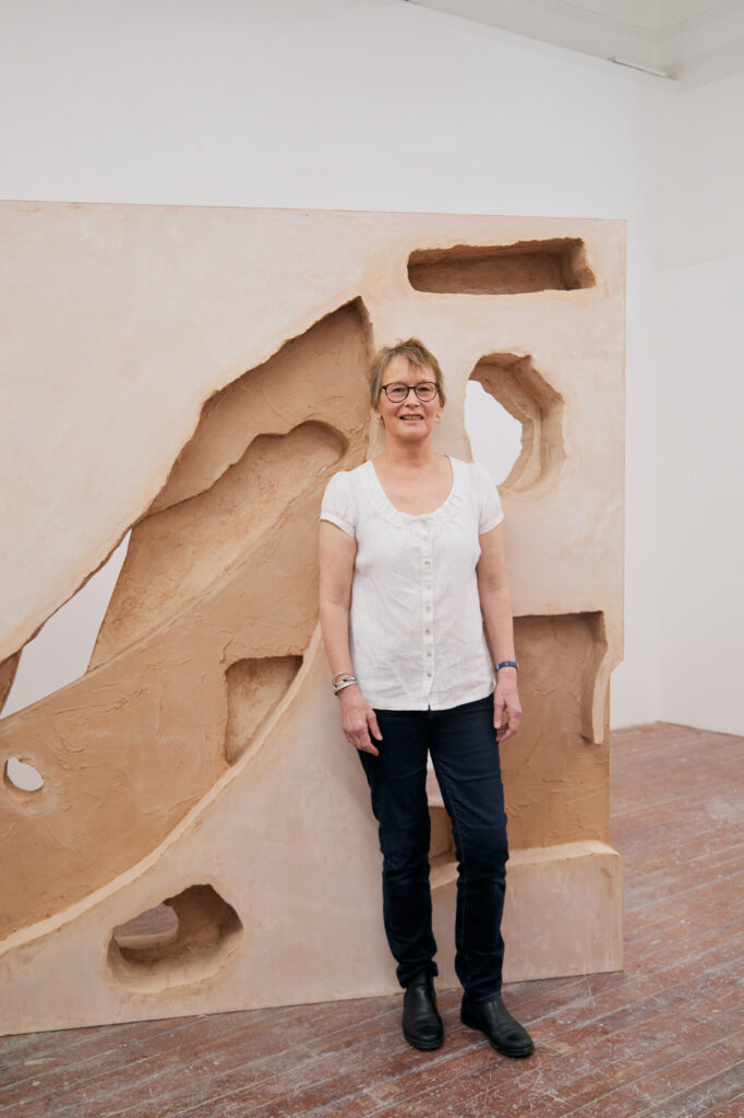 A woman with short blond hair, glasses, and a white shirt stands smiling in front of a large beige sculptural artwork with geometric cutouts, on a wooden floor in a bright gallery space.