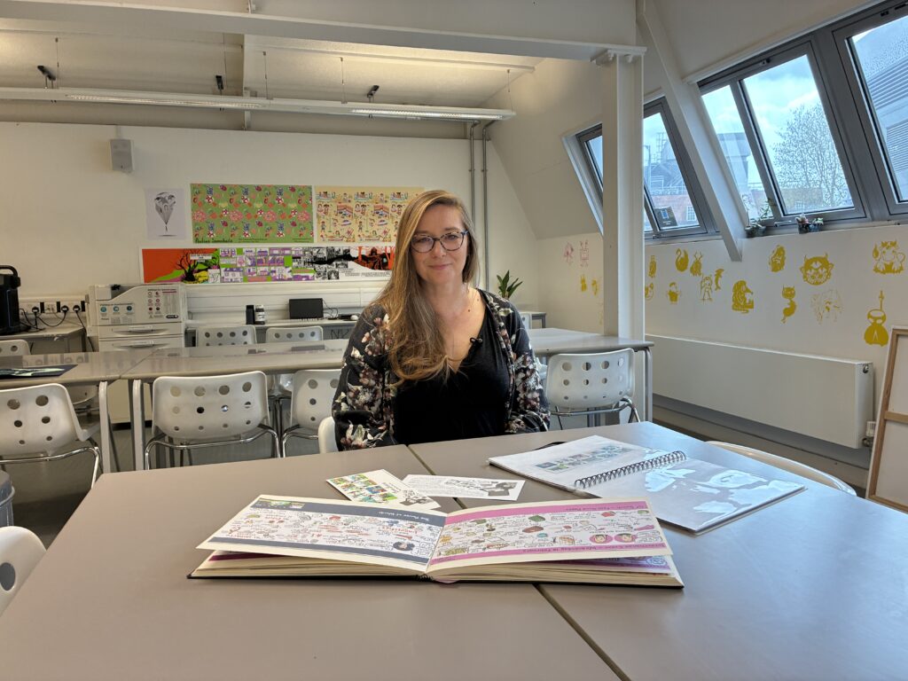 A woman with long hair and glasses sits at a table in a bright art classroom, surrounded by sketchbooks and drawings. colourful artwork is displayed on the walls and sunlight streams in through large windows.