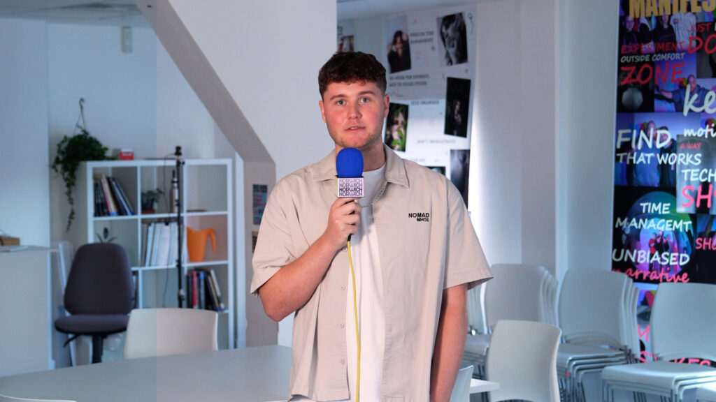 A young man stands indoors holding a blue microphone, facing the camera. Behind him are white shelves with books, chairs, and colorful posters on the wall. The setting appears to be a creative or office space.