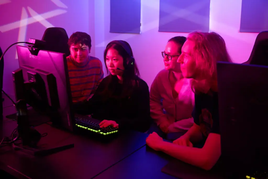 Four people sit together at a desk in a room with purple lighting, focused on a computer monitor. One person wears a headset and uses the keyboard and mouse while the others watch attentively.