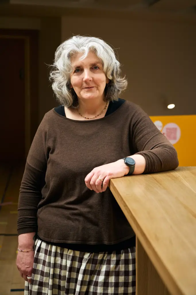A woman with gray curly hair, wearing a brown top, checkered skirt, and a black watch, stands indoors leaning on a wooden counter, looking at the camera with a neutral expression.