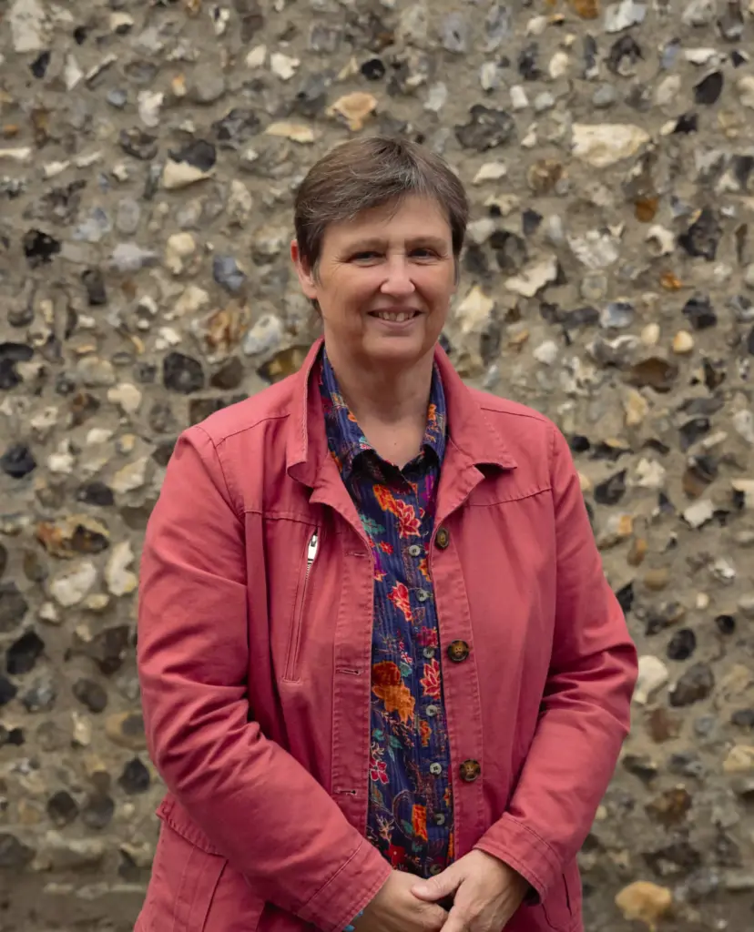 A person with short hair wearing a coral jacket over a patterned shirt stands in front of a stone wall, smiling with hands clasped in front.