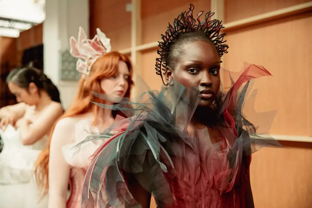 Three models stand in line backstage. The front model wears a dramatic dark tulle dress with spiky details. Behind her, models with long hair wear pastel dresses, one adjusting her outfit. Shelves and wood panels are in the background.
