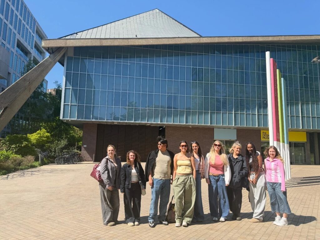 A group of nine people stand in a row outside a modern building with large glass windows and a slanted roof, smiling in the sunlight. Most are wearing casual clothes, including jeans and jackets.