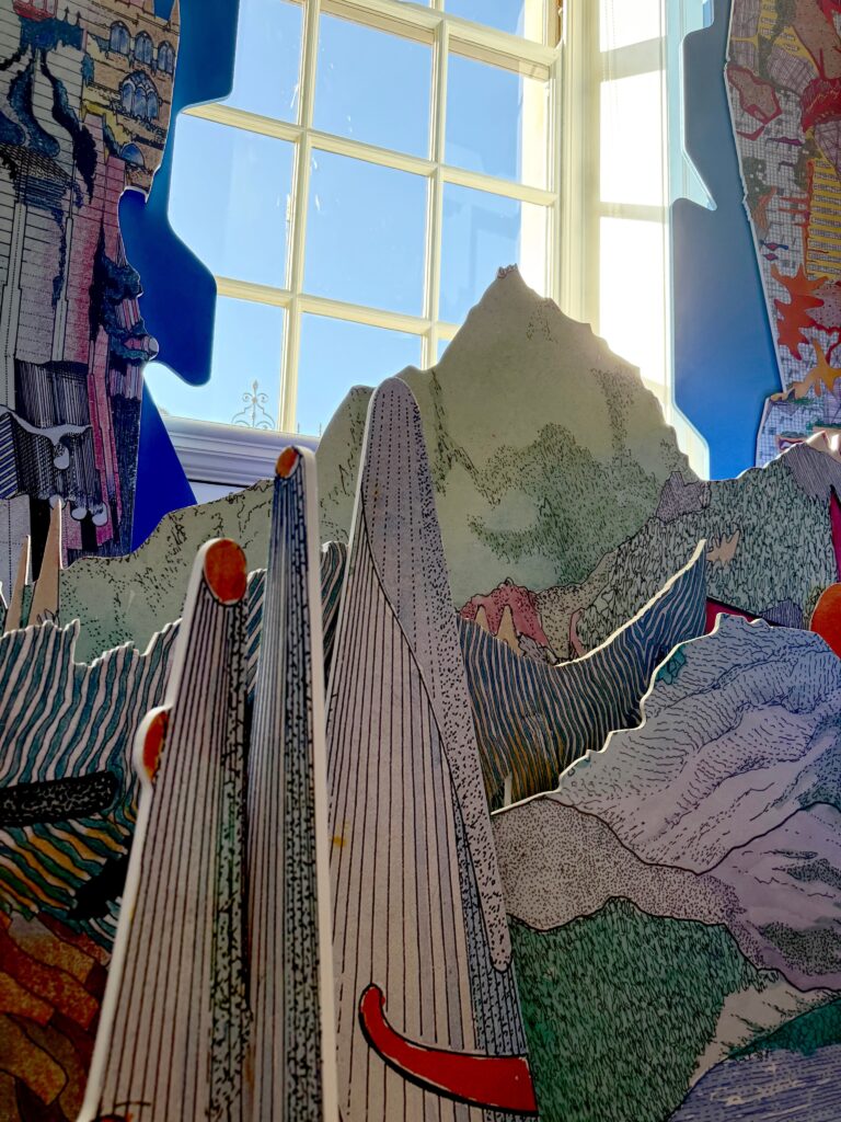 Colorful, abstract cardboard cutouts resembling mountains and landscapes are arranged in front of a bright window with a grid pattern, letting sunlight into the scene. The cutouts have fine, detailed line work and vibrant colors.