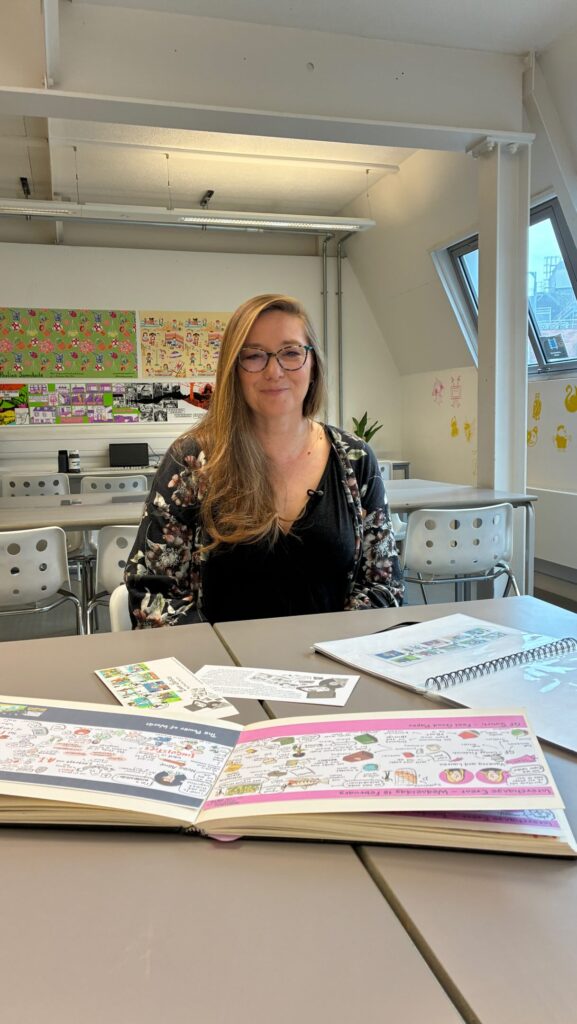 A woman with long hair and glasses sits at a table in a bright classroom, smiling. Open sketchbooks with colorful drawings and papers are spread out in front of her. Artwork decorates the back wall.