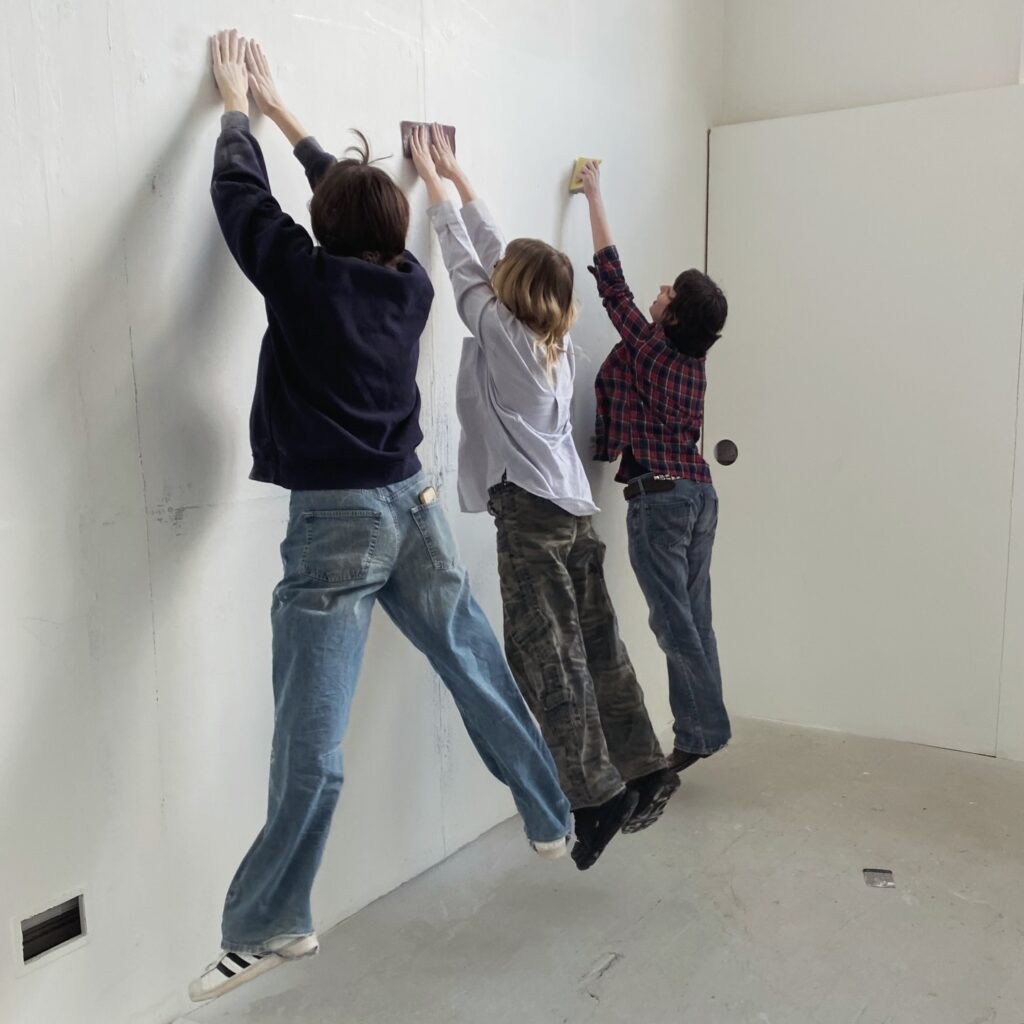 Three people are jumping and reaching up to clean a white wall with sponges in a minimalist room, dressed casually in jeans and shirts.