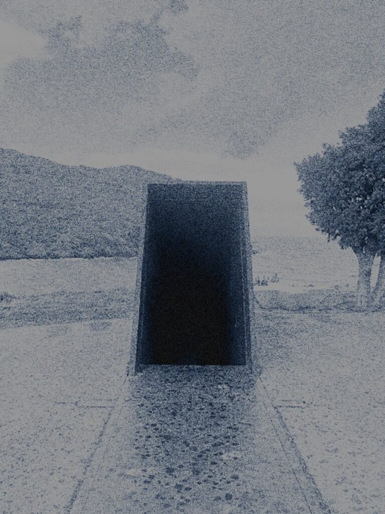 A grainy, high-contrast photo shows a dark rectangular tunnel or monument facing a body of water, with mountains in the background and a tree on the right. The scene appears stark and somewhat abstract.