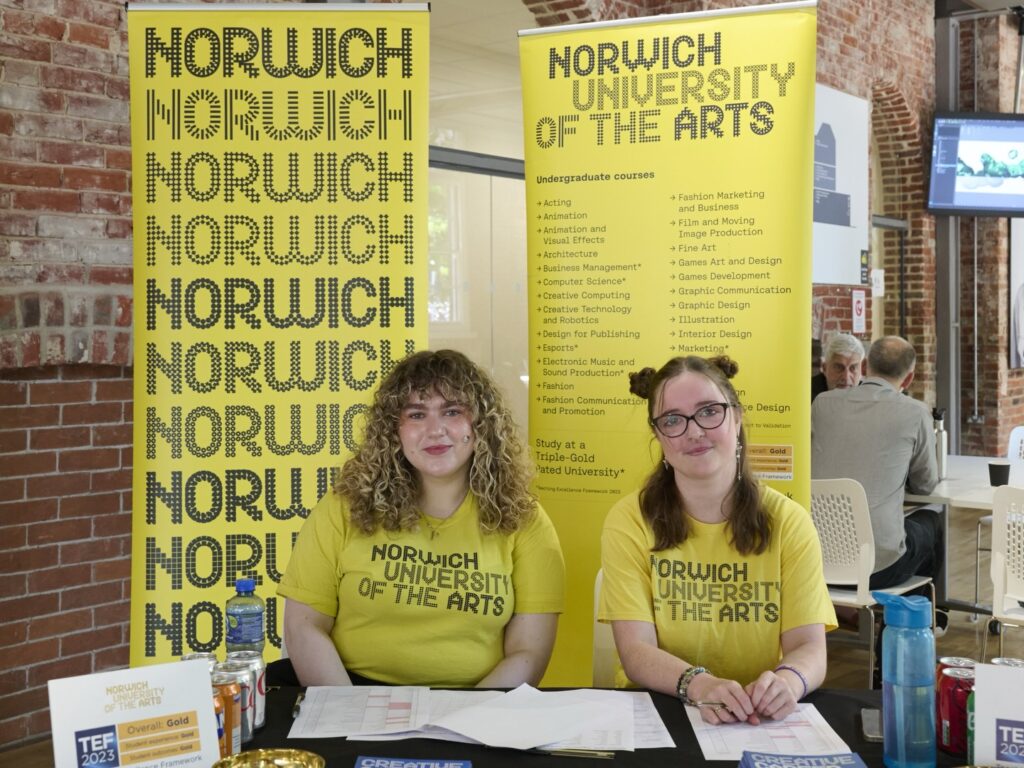 Two women sit at a table with brochures, wearing yellow "Norwich University of the Arts" shirts. Yellow banners with course information and the university name are displayed behind them. The setting appears to be an open day or event.