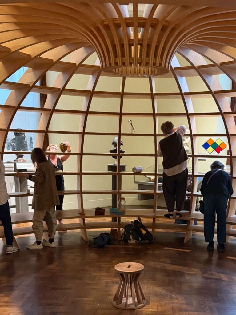 People observe and interact with various objects inside a large, wooden, ribbed dome structure in a museum or gallery. A colorful geometric artwork and sculptures are displayed beyond the wooden framework.
