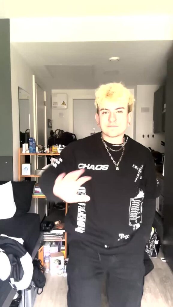 A person with short bleached blond hair wearing a black sweater with white text and chains stands smiling in a well-lit, cluttered room, waving with one hand. The room has shelves, a bed, and various personal items.
