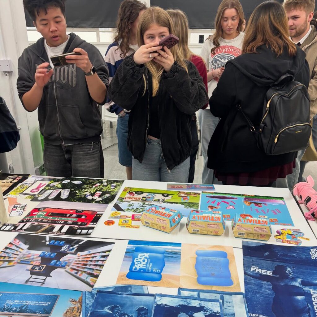 A group of young people stand around a table displaying colorful artwork and product packaging, with some taking photos of the table with their phones.
