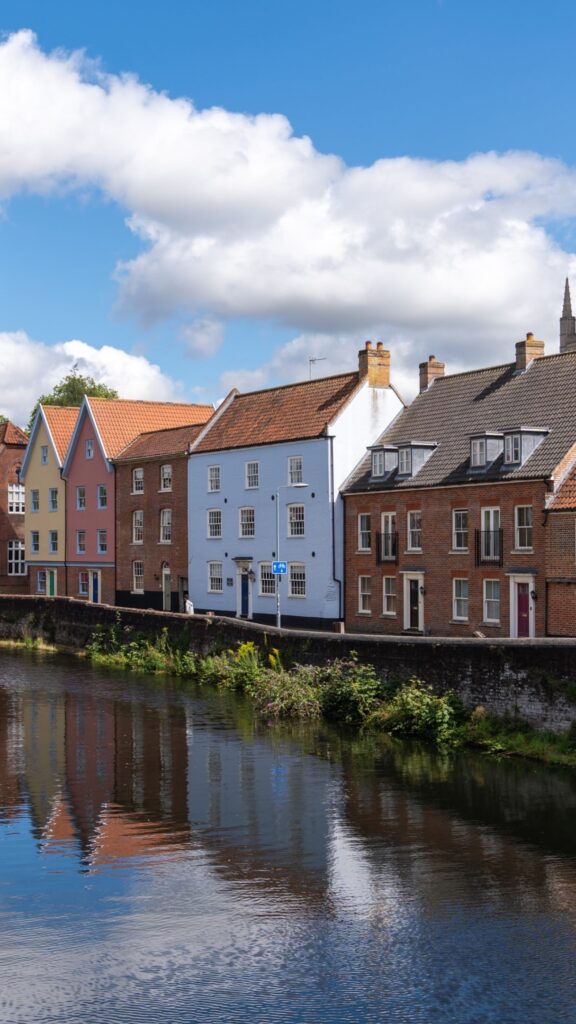 Row of colorful houses with brick roofs reflected in a calm river under a bright blue sky with scattered clouds. Some greenery grows along the riverbank.