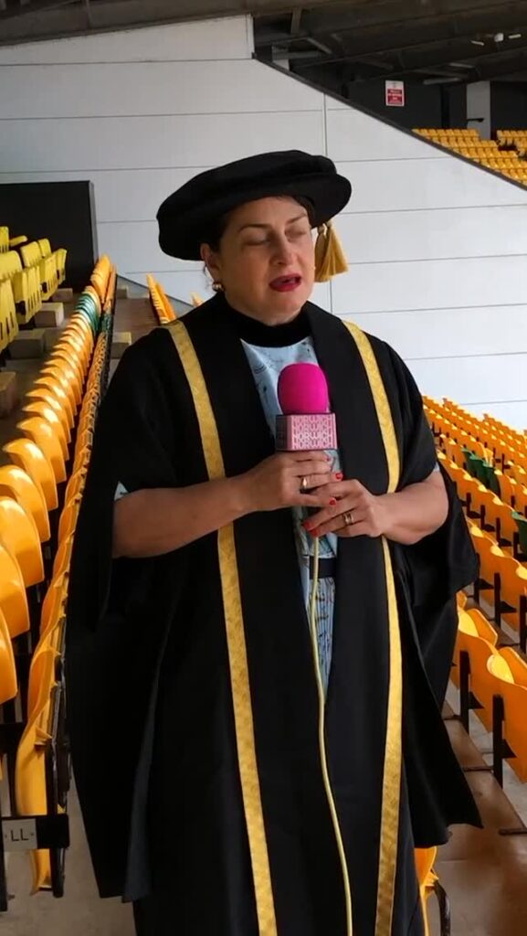 A person in academic regalia with a black robe and cap holds a pink microphone, standing near yellow stadium seats, appearing to speak or sing.