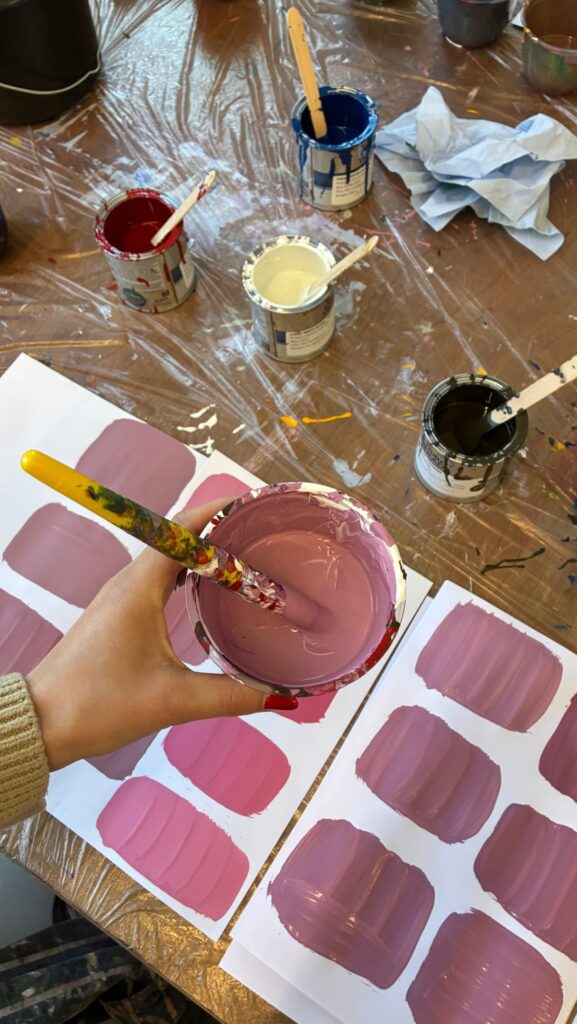 A hand holds a cup of mauve paint with a brush above a table covered with paint cans, brushes, and paper sheets showing swatches of various pink and purple shades.