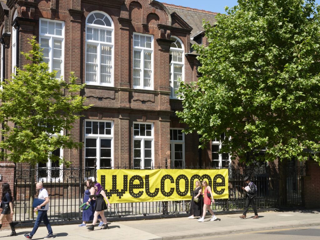 A large yellow banner with the word “welcome” in bold black letters hangs on the fence outside a brick building. Several people walk along the sidewalk in front, and leafy green trees partially shade the scene.