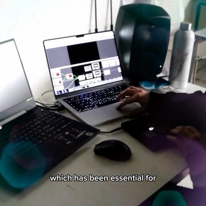 A person uses a laptop with audio-visual editing software open, next to another laptop, a computer mouse, a large speaker, and a water bottle on a desk. Subtitles at the bottom read, "which has been essential for".