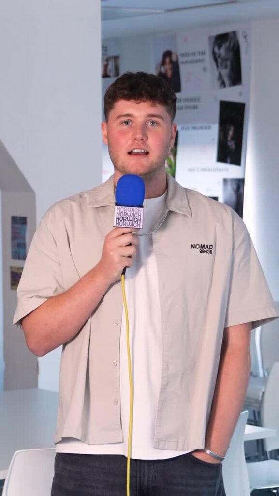Young man in a beige button-up shirt and black jeans holds a microphone with "NORWICH" logo. He has short brown hair and is looking at the camera.