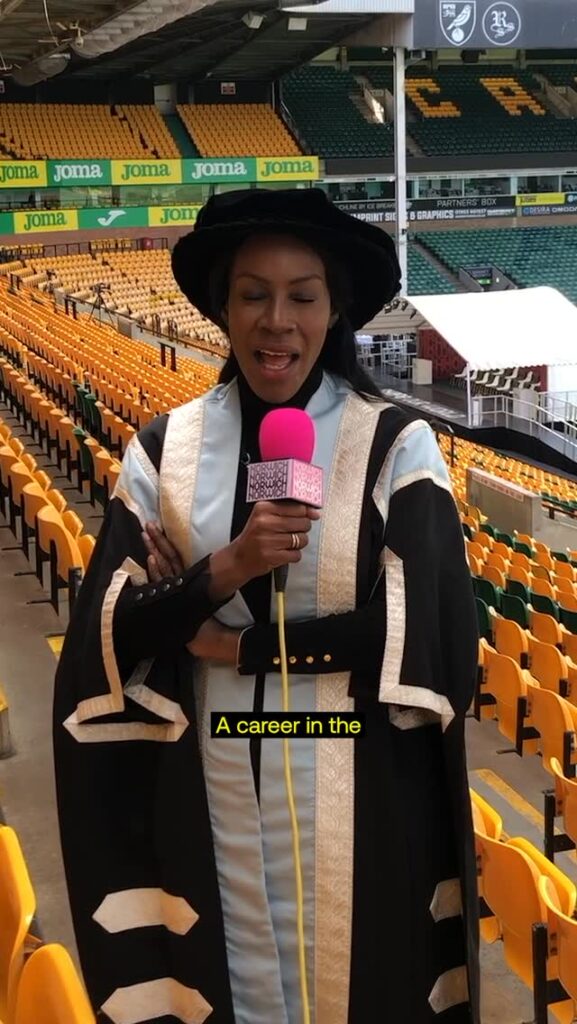 A woman in ceremonial academic attire stands in a stadium, holding a pink microphone and speaking. She is surrounded by rows of yellow seats, and the stadium appears empty.