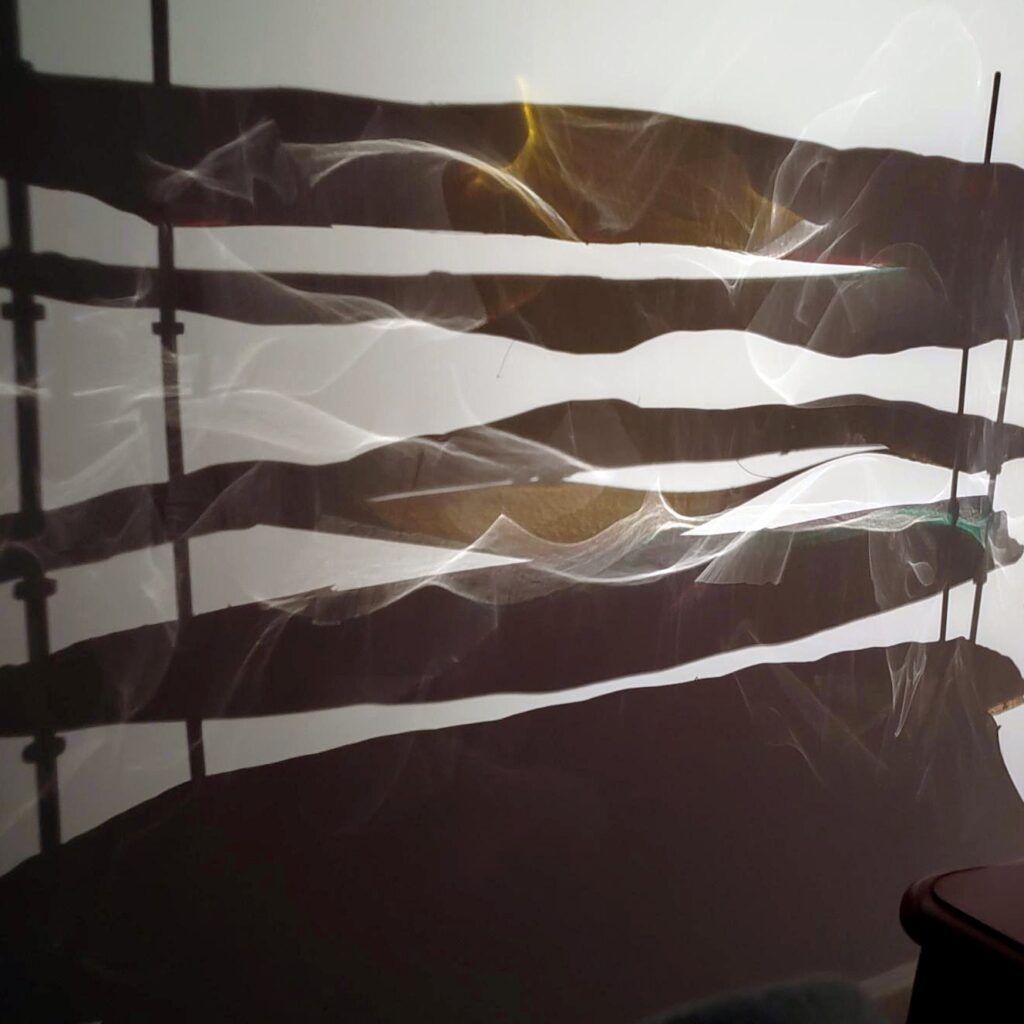 Abstract shadows and light patterns are cast on a wall by several horizontal objects, possibly fabric or slats, with wispy, flowing reflections creating an artistic effect.