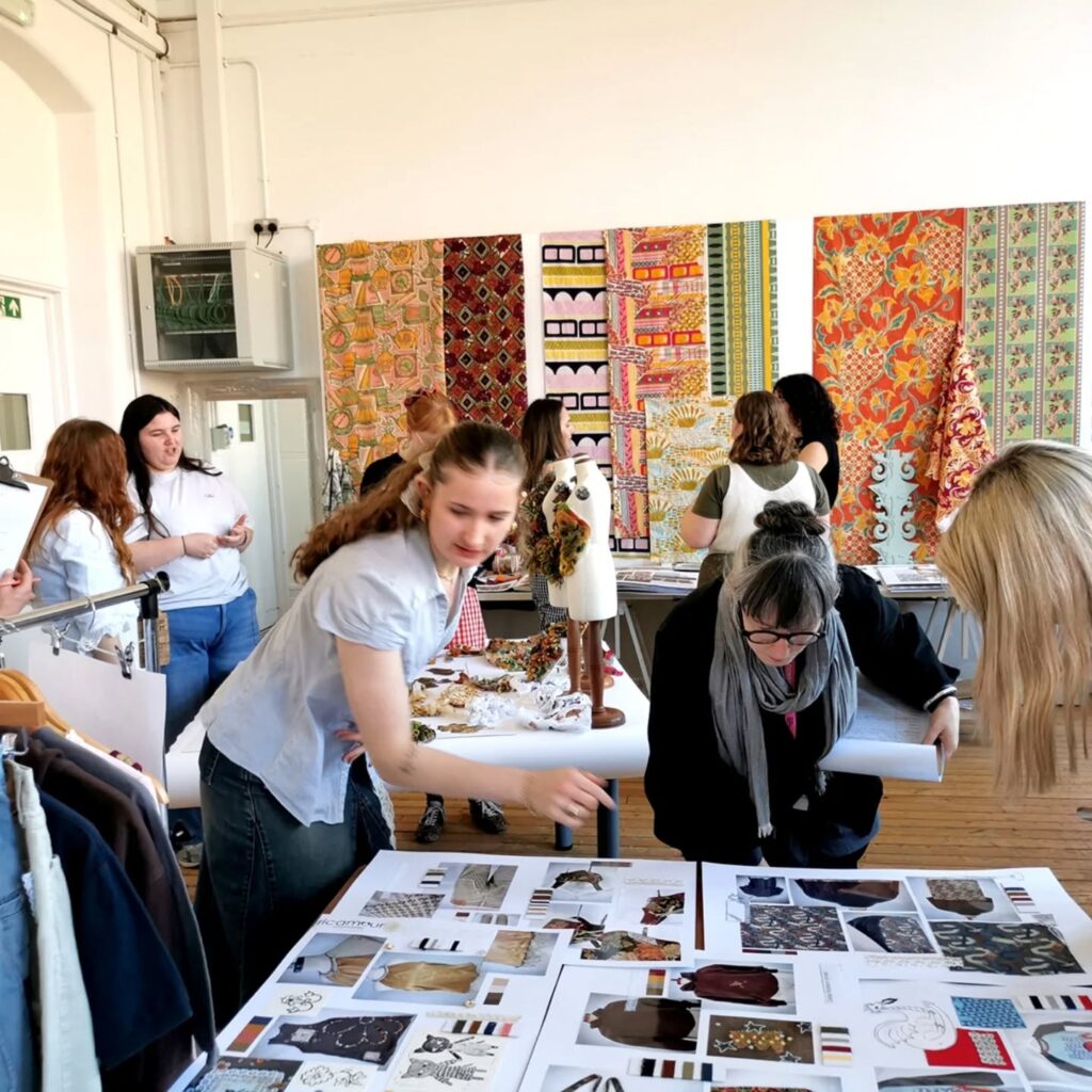 A group of people in a bright studio review fashion sketches and fabric samples on tables. Colorful patterned textiles hang on the wall behind them, and racks of clothing are visible on the left.
