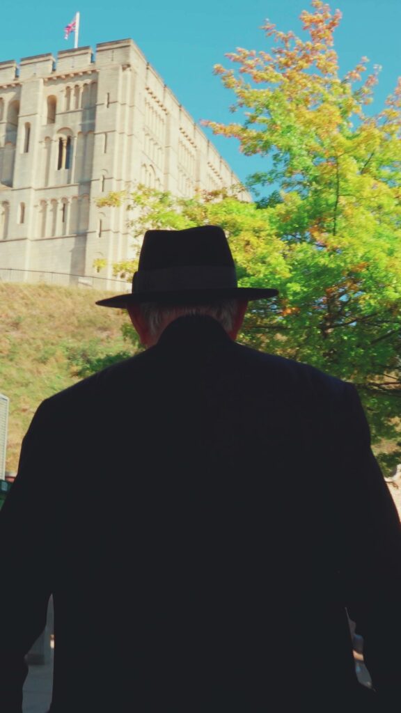 A man in a black hat and dark coat stands with his back to the camera, facing a large stone building on a sunny day, with green and yellow trees beside him.