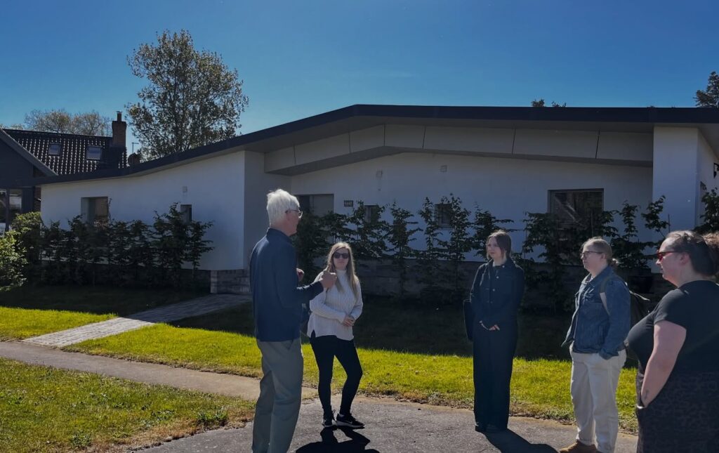 Five people stand and talk on a sidewalk in front of a modern, white, single-story house on a sunny day. The sky is clear and blue, and shadows fall on the grass and pavement.