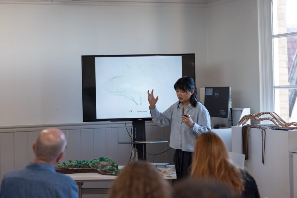 A woman stands in front of a screen with a map, gesturing as she presents to a seated audience in a bright room. There is a model with greenery on the table in front of her.