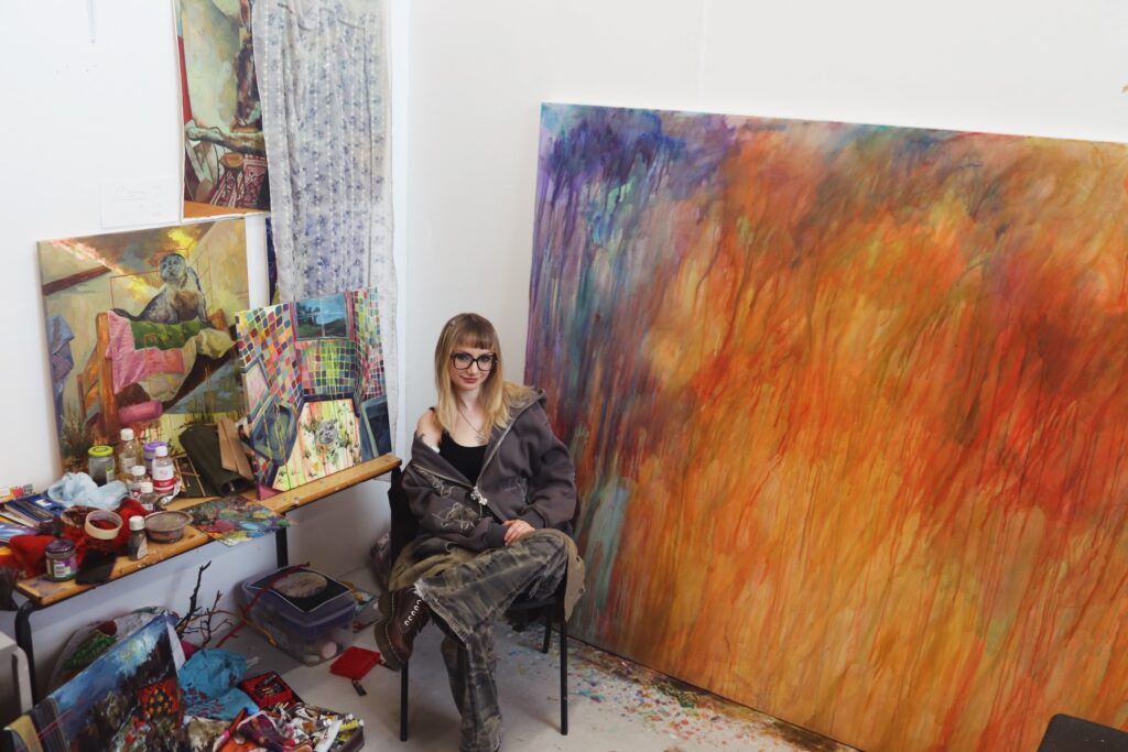 A person with long blonde hair and glasses sits in an artist’s studio, surrounded by colorful abstract paintings, art supplies, and a large, vibrant canvas with red and orange hues propped against the wall.